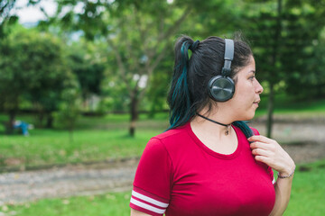 beautiful college woman with blue hair, red dress and blue backpack, listening to music with her wireless headphones in a park, girl using music to get away from the outside world, relax and think.