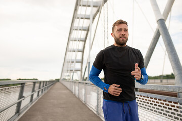 Handsome young man running in the city wearing blue sports outfit and his smart watch to check his pulse during his run. Male athlete looking to the side running on a bridge training for a marathon.