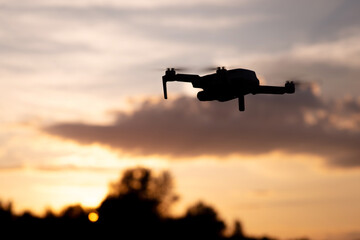 Dark silhouette of drone flying in sunset landscape. Sun behind tops of the trees, lightened clouds on the sky.