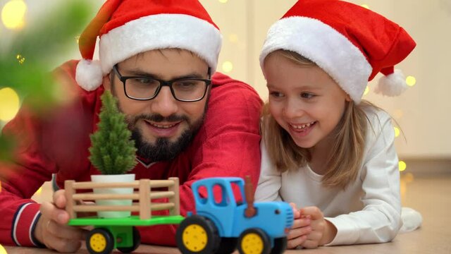 Toy tractor with cart delivers Christmas tree to father and little girl in red santa hats. Happy family with new year gift.