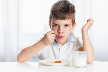Cute child in white shirt eats cottage cheese pie and drinks milk for breakfast at home