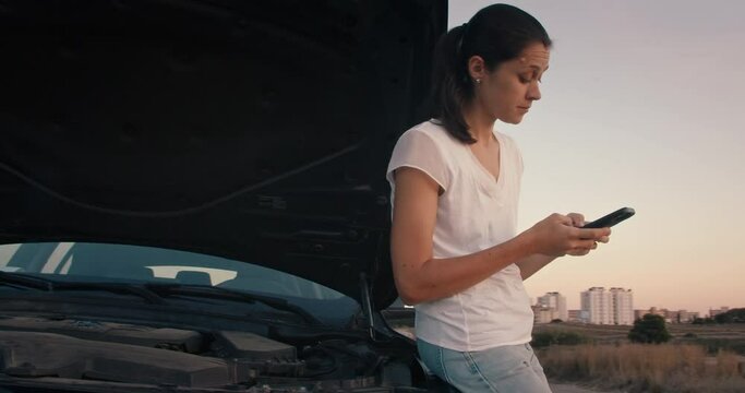 Worried woman standing front to the broken car with opened hood and using smartphone to find service. Insurance assistant on road attractive girl calls for a tow truck on sunset 