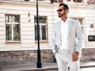 Fototapeta premium Portrait of handsome confident stylish hipster lambersexual model.Sexy modern man dressed in elegant white suit. Fashion male posing in the street background in Europe city at sunset. In sunglasses