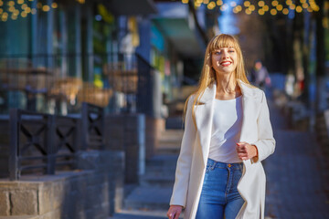 Pretty trendy girl in white coat walking on street. Business woman rests on the city. Fashion woman close-up portrait, autumn or spring, laughing and smiling young model. Blurred details.