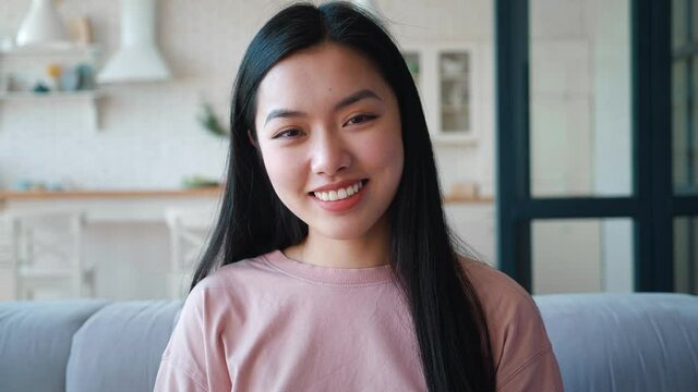 Portrait of confident beautiful young Asian woman sitting at couch at home. Charming young Asian ethnicity woman smiling with toothy smile, looking confidently into the camera, sitting on sofa at home