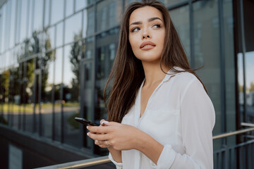 Attractive girl working in office corporation, company, dressed in elegant clothes, stands leaning against railing in front of glass building, holding phone in hands, waiting for call, looking to side