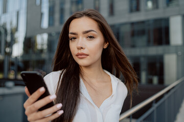A pretty young woman with serious face employed in a corporate office walks out of a glassy modern building outside for a break, holding phone in hand, reading messages, writing back to friends