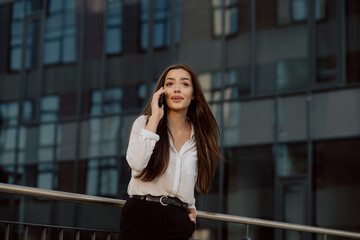 Beautiful girl stands leaning against railing in front of a modern glass office building, businesswoman dressed in a white shirt holds the phone to ear, talking to friend, smiles surprised face