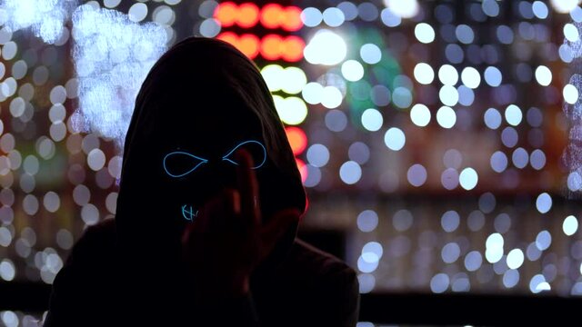 A masked man with glowing eyes and teeth in the image of death beckons with a hand gesture to himself. It stands against the background of neon lights in the dark