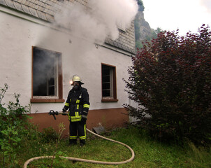 Feuerwehrmann steht mit Schlauchleitung an einem Fenster einer brennenden Wohnung