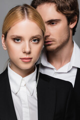 elegant blonde woman looking at camera near blurred young man isolated on grey