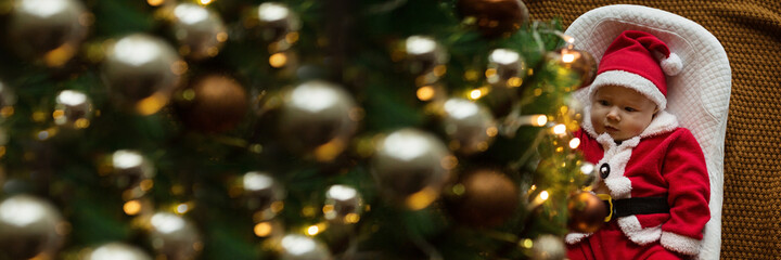Closeup portrait of newborn baby. Cute Caucasian baby girl 4-5 months old in Santa costume lying on knitted cozy blanket in cocoon near decorated fir tree. Merry Christmas xmas and happy new year 2022