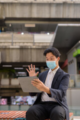 Asian businessman wearing face mask and sitting outdoors in city using digital tablet computer