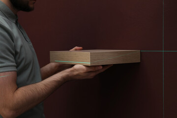 Man using cross line laser level for hanging shelf on brown wall, closeup