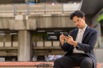 Asian businessman sitting outdoors in Bangkok city using mobile phone