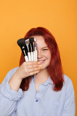 Portrait of young smiling visagist with makeup brushes in hand. Red-haired girl isolated on orange. Professional