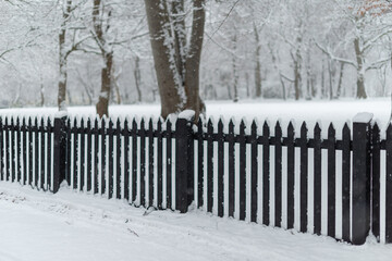 A dark wood picket fence separating a road and field covered in snow. The field is part of a park...