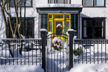 The entrance to a white house with a yellow wood and glass door. There's a black metal wrought iron fence and gate in the garden. A Christmas wreath hangs from it. The ground is covered in white snow. - Powered by Adobe