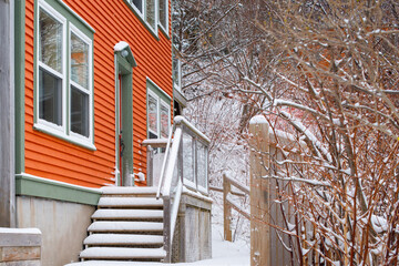The exterior facade of an orange color wooden house with green and white trim. There are steps leading up to the entrance covered in white snow. It is a winter scene of a traditional wood house.