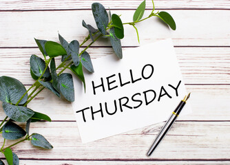 On a light wooden table, there is a eucalyptus branch, a fountain pen and a sheet of paper with the text HELLO THURSDAY