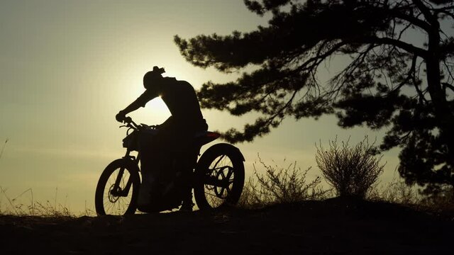 A silhouette of young man with beard and glasses sitting on motorcycle at summer sunset. Man leaves his bike and follows the sunshine. Evening at pine forest, non urban scene
