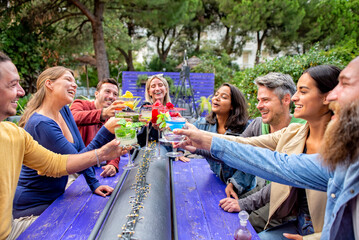 happy diverse large group of multicultural friends making celebratory toast holding beautiful gourmet cocktail glasses sitting at bar restaurant outdoor. convivial people having fun. lifestyle concept