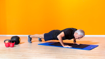 focused senior fit man doing push up workout exercise laying at the gym isolated on orange background. mature people fitness training indoor. elderly person recovery healthy lifestyle concept
