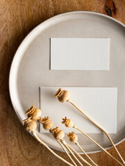 Blank greeting card, invitation and dried flowers on beige ceramic plate and round wooden tray in morning sunlight, flat lay, top view