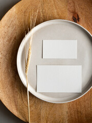 Blank greeting card, invitation and dried flowers on beige ceramic plate and round wooden tray in morning sunlight, flat lay, top view
