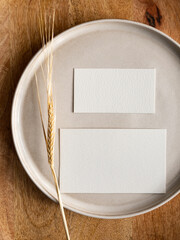 Blank greeting card, invitation and dried flowers on beige ceramic plate and round wooden tray in morning sunlight, flat lay, top view