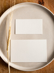 Blank greeting card, invitation and dried flowers on beige ceramic plate and round wooden tray in morning sunlight, flat lay, top view