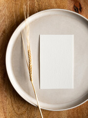 Blank greeting card, invitation and dried flowers on beige ceramic plate and round wooden tray in morning sunlight, flat lay, top view
