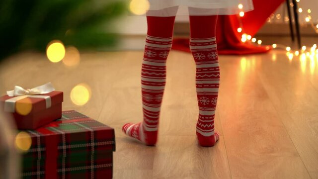 Little childrens feet in red tights with New Years ornament dance next to Christmas tree with gifts. Christmas socks