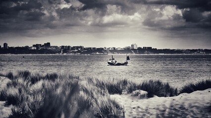 Fishing boat sailing into Poole Harbour with Bournemouth in the background, and Shell Bay sand dunes in the foreground, black and white sepia