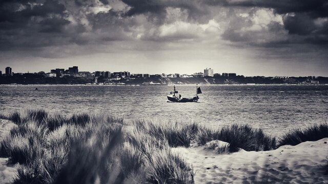 Fishing boat sailing into Poole Harbour with Bournemouth in the background, and Shell Bay sand dunes in the foreground, black and white sepia