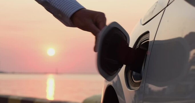 A man's hand pulls out the charging plug of an electric car from the car charging socket against the backdrop of the rising sun. Dawn of the era of electric transport.