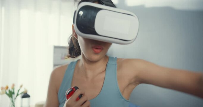 Young attractive Asian woman with sportswear wearing virtual reality headset play switch game boxing exercise online program in living room at house. Home quarantine workout and fitness concept.
