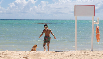 BOY PLAYING WITH HIS DOG IN THE BEACH SAND, 9-YEAR-OLD  IN THE SHORES OF A TURQUOISE BLUE CARIBBEAN BEACH WITH CLEAR SKY AND BLANK SIGN