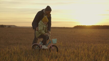 father teaches little kid to ride a two-wheeled bike in the sunshine, happy family, fun childhood life, child and dad together on the weekend have fun on a bike, play a game with parent at sunset