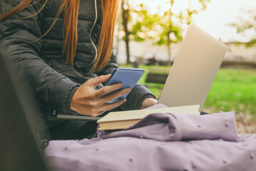 Girl study sitting on a bench in the park in a beautiful autumn afternoon. Teen using laptop, smartphone and reading a book outdoor. Student alone doing homework outside in the nature. School  concept