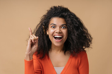 Young excited African American woman pointing finger up having idea, good news. Emotional curly haired female with open mouth looking at camera isolated on beige background. Inspiration concept 