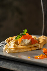 Crepes (big thin pancakes) with red caviar, and cream cheese or sour cream with basil leaf on dark background. Beautiful light, selective focus, copy space. Tasty breakfast, dark mood photo concept.