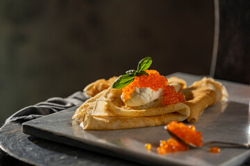 Crepes (big thin pancakes) with red caviar, and cream cheese or sour cream with basil leaf on dark background. Beautiful light, selective focus, copy space. Tasty breakfast, dark mood photo concept.