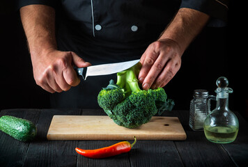 Professional chef cuts fresh Broccoli with a knife for salad on vintage kitchen table with fresh vegetables. Cooking and restaurant or cafe concept.
