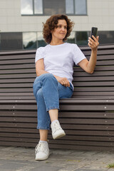 A 45-50-year-old woman is sitting on a bench and taking a selfie on a mobile phone