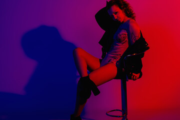 Fashion model in stylish clothes at the neon lights in the studio. Studio portrait of a sexy woman in a leather jacket, bodysuit and boots on a red, blue, purple ultraviolet background in a nightclub