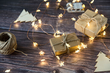 Fototapeta na wymiar Glowing garland star, two gray boxes with gifts tied with twine, plywood Christmas decorations carved from wood on a wooden table surface