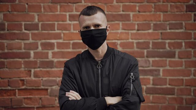 Caucasian gloomy male security guard in a black jacket against the background of a brick wall looks into the camera, removes the protective mask, spits to the side