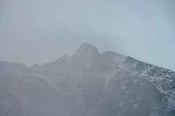 Fototapeta na wymiar Wonderful view to mountain silhouette with pointed peak in fog. Atmospheric foggy mountain landscape with snow and peaked top in low clouds. Minimalist alpine scenery with sharp rocky pinnacle in mist
