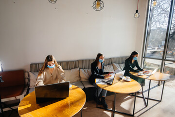 A group of masked girls keep a social distance in a cafe when working on laptops.